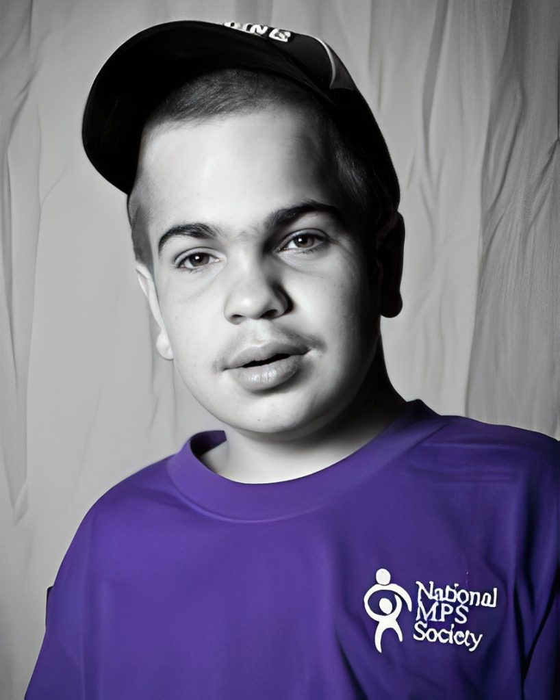 869ae4dc2b23c35b96325aeb6e6f353d Person wearing a baseball cap and a purple National MPS Society shirt, standing in front of a plain backdrop.