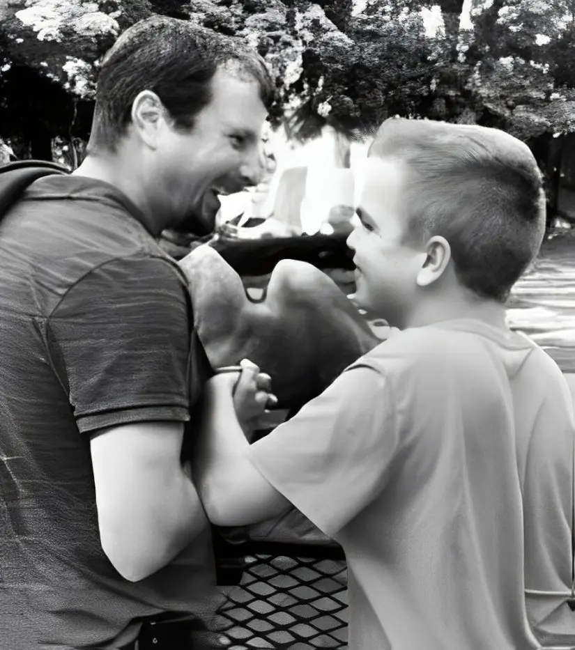 ed621791eaf0e405b575aebcdb4956be Adult and child smiling and shaking hands in an outdoor setting with trees in the background.